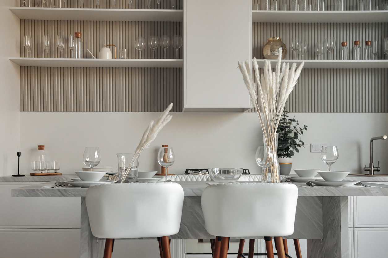 Modern interior of home white kitchen room with design furniture - served table, shelves and elegant personal accessories. Stylish neutral domestic decor. Designer style concept.. Contemporary modern kitchen with white and wood bar stools and white and glass kitchen accessories.